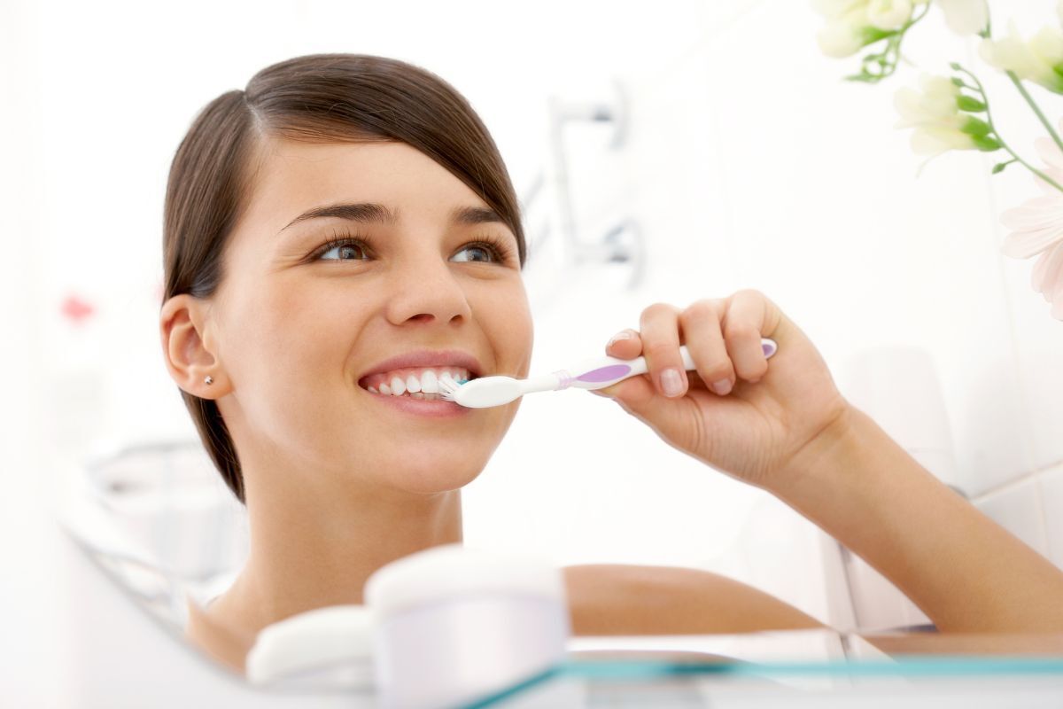 5 Tips for Better Oral Hygiene Woman brushing her teeth with a purple toothbrush in a bright bathroom setting.