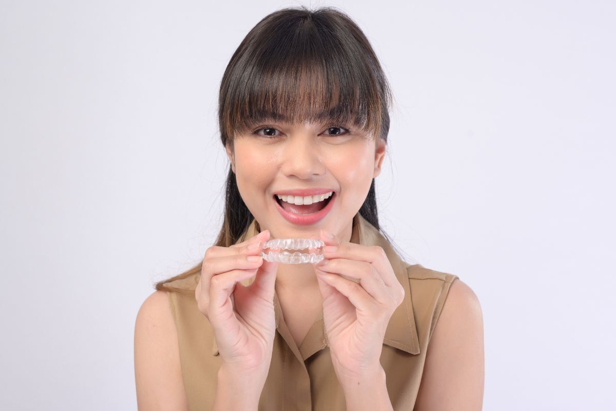 3 Types of Invisible Orthodontics Young woman smiling while holding an invisible aligner tray for orthodontic treatment.
